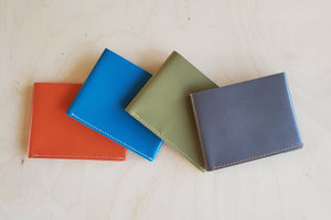 Simple Flap wallets in orange, blue, grey and olive green leather and white stitching from architect Alice Park shown folded.