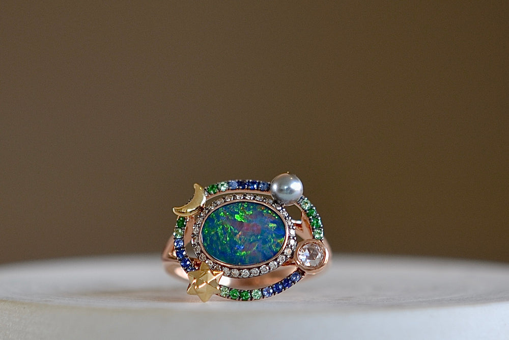 Small Galaxy Opal Ring by Bibi Van Der Velden is a rose gold ring with an opal, surrounded by a ring of brown pave diamonds, another ring of blue sapphires and green pave tsavorites, a gray keshi pearl, a white rose cut diamond, a star and moon in yellow gold. 
