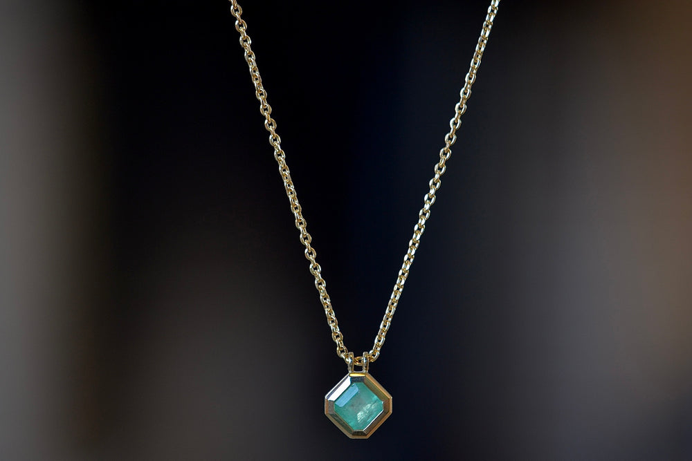 The Duo Bale Offset Emerald Necklace by Elizabeth Street is a bezel set, translucent and rectangular shaped Columbian emerald hanging on an axis with twin bales and on a 14k yellow gold chain. 