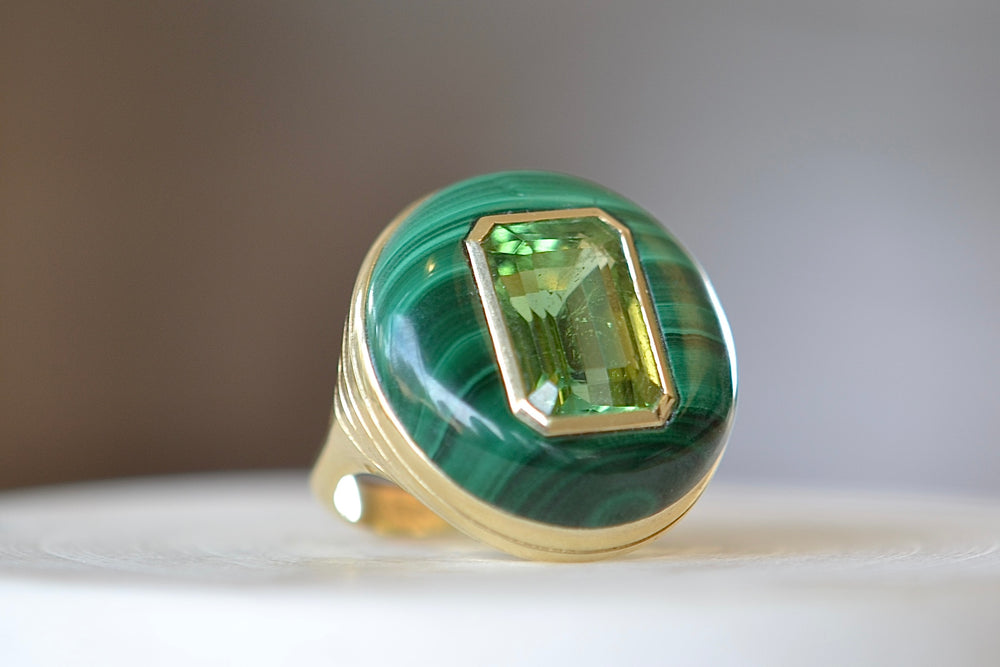 The Large Lollipop Ring in malachite and Green tourmaline by Retrouvai is a one of a kind signet ring featuring a chunky and rounded stone face with a translucent center stone set on a tiered edge and tapered band. This one is malachite with an emerald cut green tourmaline in the center. 