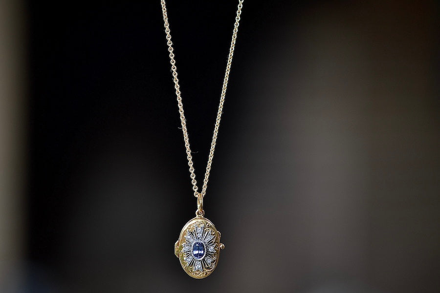 A Blue Sapphire Locket that opens by Arman Sarkisyan is an engraved oval disc in 22k with a blue center stone inside a flower motif made out of diamonds and hangs on a gold chain. 