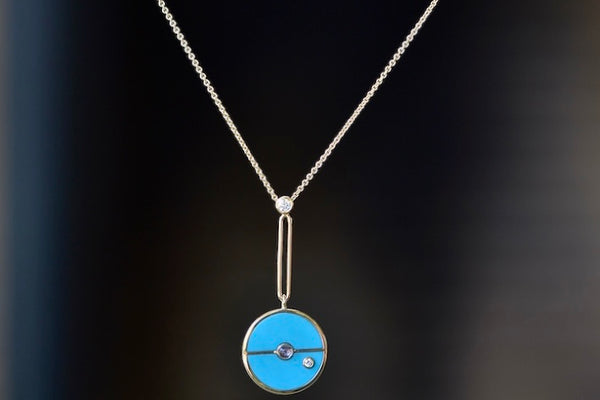  The signature Compass Pendant with Sleeping Beauty Turquoise and Tanzanite by Retrouvai is a pendant necklace with stone inlay that is accented with a center gem stone and a white diamond. The compass hangs from an elongated paperclip link accented with a second white diamond on 24" 14k yellow gold chain.