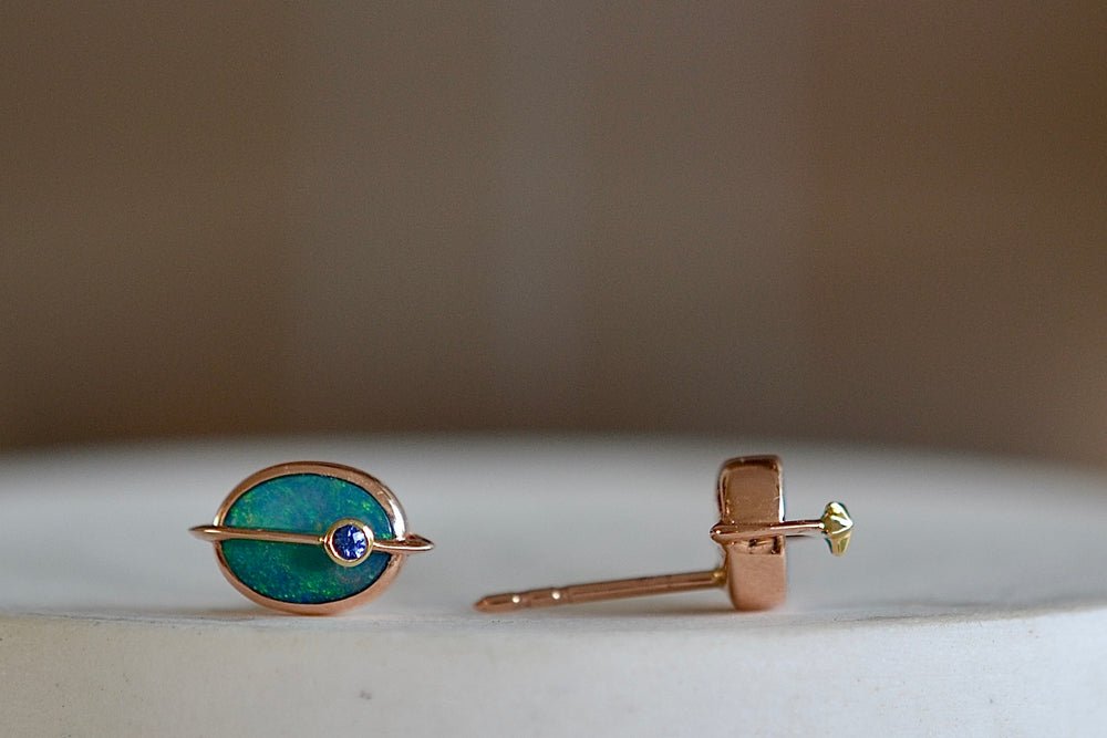 Side view of Small Planet Stud Earrings by Bibi Van Der Velden are comprised out of two bezel set opals with rings going through in rose gold of which one has a gold star and the other has a blue sapphire planet. 
