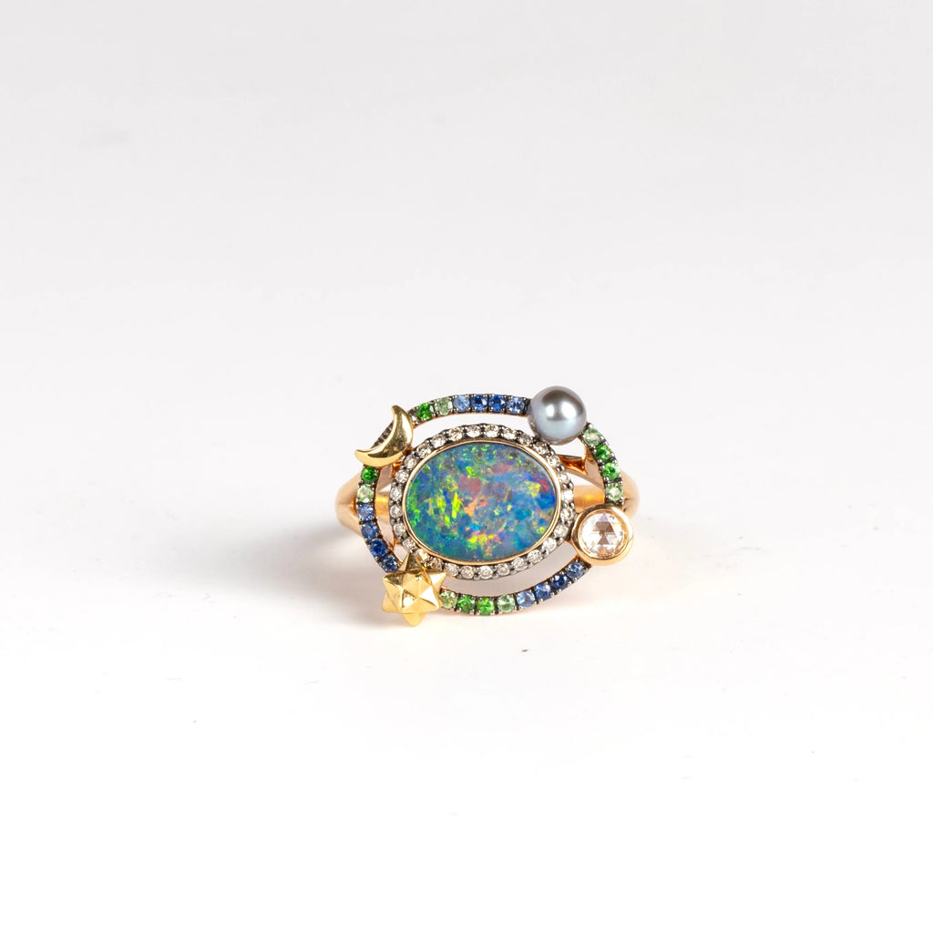 Small Galaxy Opal Ring by Bibi Van Der Velden is a rose gold ring with an opal, surrounded by a ring of brown pave diamonds, another ring of blue sapphires and green pave tsavorites, a gray keshi pearl, a white rose cut diamond, a star and moon in yellow gold. 
