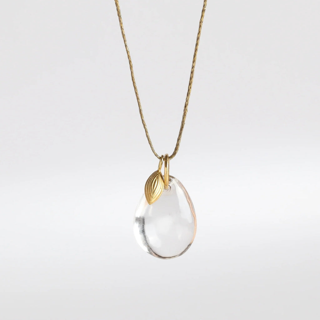 The Coconut Pendant with Seed Charm Necklace in Crystal  designed by Pippa Small is a One clear, transparent and smooth crystal stone with natural inclusions is drilled and hooked on to a gold loop, and accompanied by a bead in 18k yellow gold on a 24" golden waxed cotton cord form this necklace.