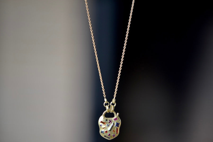 The Small Harlequin Coeur de Confetti Padlock Necklace by Polly Wales is a padlock in 18K yellow gold that clicks open on the side and has scattered rainbow sapphires in pink, blue, red, yellow and orange and hangs on a beautiful chain. The chain attaches to the lock handle by pulling the lever.  