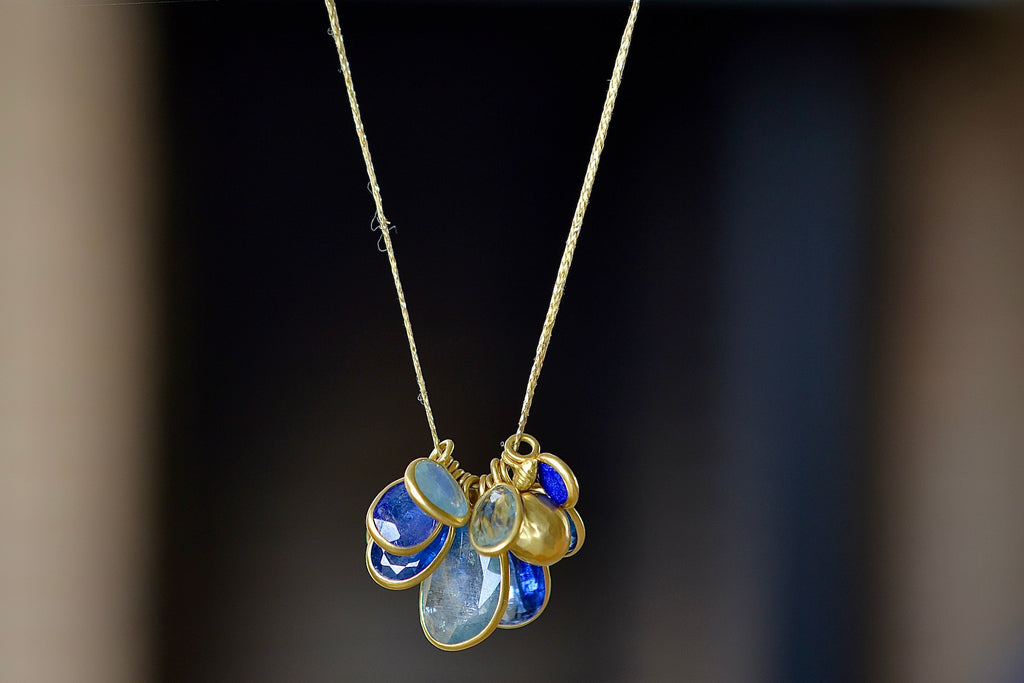 The Colette Mixed Blues Cluster Necklace by Pippa Small is A large cluster of nine various sized, bezel set, lightly faceted and translucent stones in the peacock color-way with aquamarine, amethyst, labradorite, kyanite, lapis and tanzanite, accompanied by a hammered gold seed and mini croissant beads in 18k gold and hanging on a 16" golden waxed cotton cord.