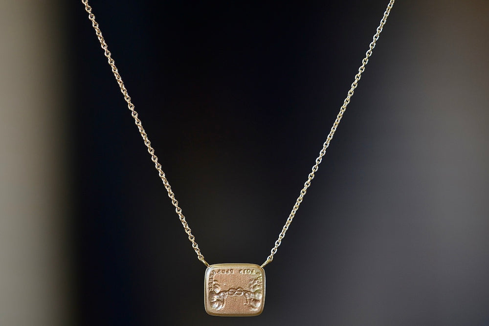 Love Knot / Peace and War Duo Motto Necklace designed by Makiko Wakita is made by by stamping an 1800s carved seal with 'mottos' in French, Italian, Latin or English on to gold. A rectangular disc in gold with satin finish that will turn shiny over time features an image of hands in the sky, each holding a piece of the tie that binds them together. Word surrounding the image in French: “Plus Loin Plus Serré” which translates to ‘the further apart the tighter the bond.’ Far apart, close at heart… You and me 