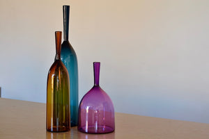 Angelic Bottles by Joe Cariati are mouth blown  Freeblown by glass master Joe Cariati in his Southern California studio, these bottle-form vases evoke the more mass produced mid-century ones that inspired them, large steel blue, small amber orange and wide ultra violet purple.