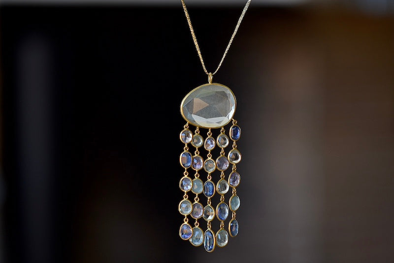 Raincloud Pendant Necklace by Pippa Small is a Large, faceted and transparent aquamarine with five rows of five stones each in blue to light purple hued tanzanite, aquamarine and kyanite bezel set stones in 18k yellow gold w extra long golden waxed cotton cord and accent bead for draping over head and no clasp.