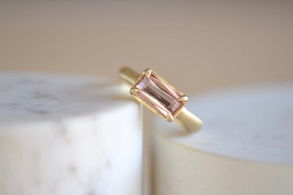 Elizabeth Street Jewelry Eagle Claw Bar ring in light pink peach tourmaline , clear and translucent, tourmaline is a radiant cut pink tourmaline in a four prong eagle claw east/west bezel setting on a 14k satin 3mm yellow gold band size 7..