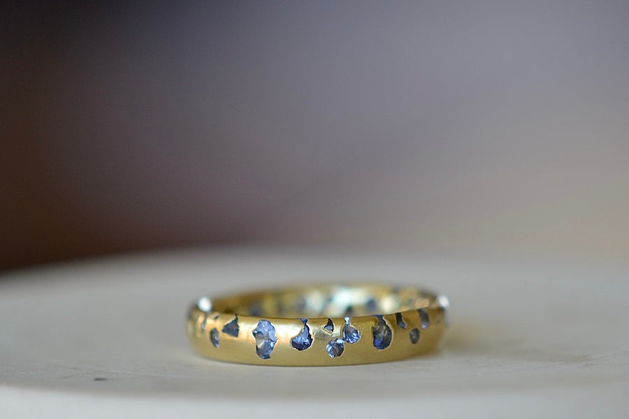 The Cornflower Blue Sapphire Confetti Band designed by Polly Wales is a narrow 18k yellow recycled gold wedding band/ring with speckled ombre light blue sapphires around the circumference. It is cast not set and handmade in Los Angeles.