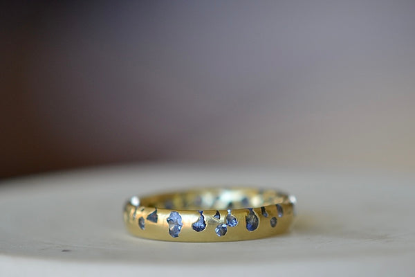 The Cornflower Blue Sapphire Confetti Band designed by Polly Wales is a narrow 18k yellow recycled gold wedding band/ring with speckled ombre light blue sapphires around the circumference. It is cast not set and handmade in Los Angeles.