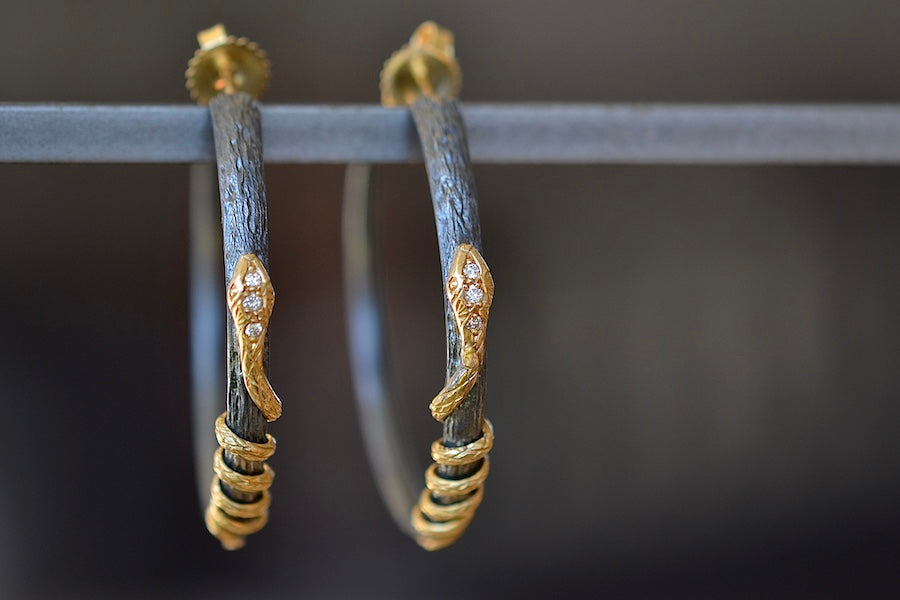 Close up of The Snake Hoop Earrings by Arman Sarkyssian are two 22k gold snakes with diamond pavé details that are entwined on an oxidized silver hoop textured like a tree branch in oxidized sterling silver with a post backing.