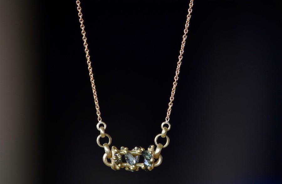 Close up of Horizontal Fontaine Bar Necklace in Green by Polly Wales is An oval bar in 18K yellow gold has a vine of encrusted inverted sapphires in green, gray and black that have been cast and are accompanied by matte gold dots. The bar hangs on a beautiful gold chain. Recycled Gold. Handmade in Los Angeles. Cast not set.