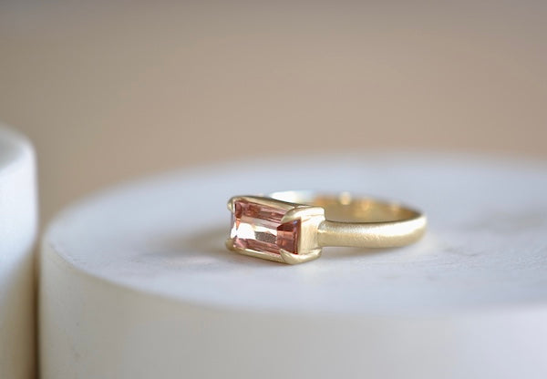 Elizabeth Street Jewelry Eagle Claw Bar ring in light pink peach tourmaline , clear and translucent, tourmaline is a radiant cut pink tourmaline in a four prong eagle claw east/west bezel setting on a 14k satin 3mm yellow gold band size 7..