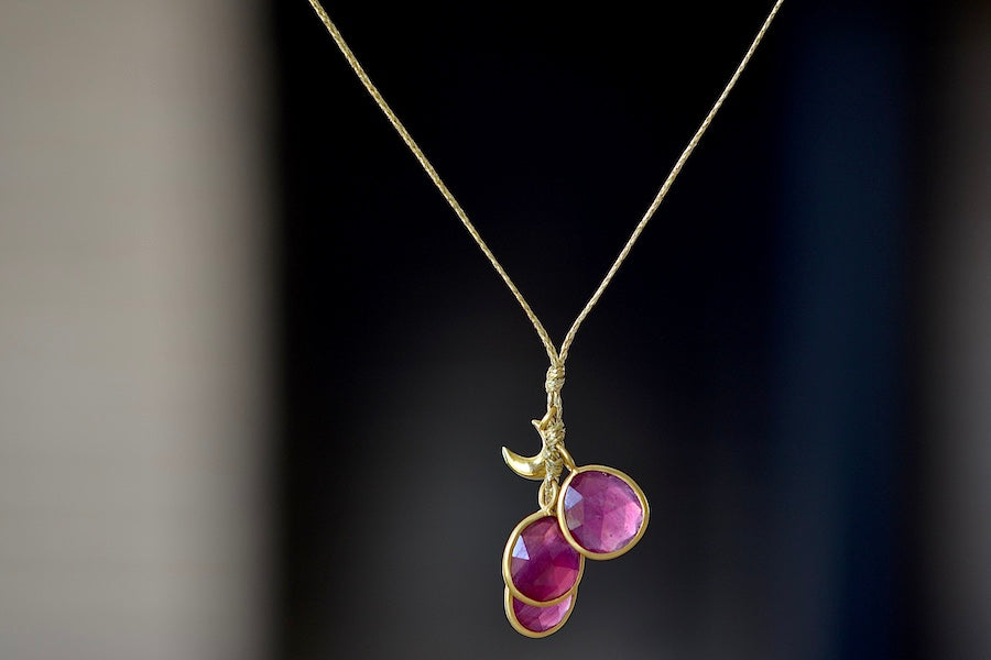The Colette Set Cluster with Moon and Ruby Necklace is a  cluster of three bezel set and lightly faceted ruby stones, accompanied by a gold moon, all in 18k yellow gold on a 22" golden waxed cotton cord form this necklace. 