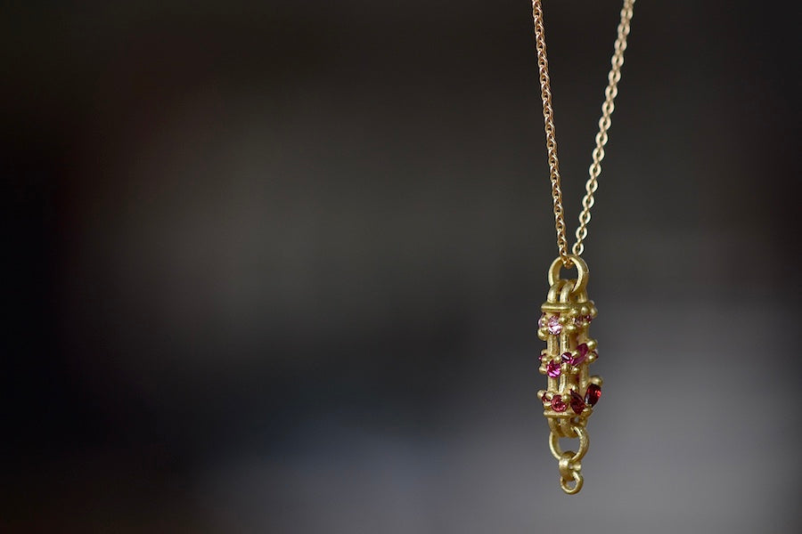 Details of stones and bar on Vertical Fontaine Bar Necklace in Plum Blossom by Polly Wales is An oval and three dimensional bar in 18K yellow gold holds a vine of encrusted and inverted sapphires in pink, fuchsia and red that have been cast and are accompanied by matte gold dots. The bar hangs on a beautiful gold chain and bale.