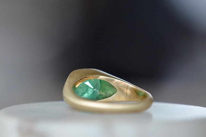 A signet ring in green emerald by Elizabeth street jewelry is a vivid cushion cut and lightly faceted Colombian emerald on a tapered band in 18k satin finish yellow gold. 
