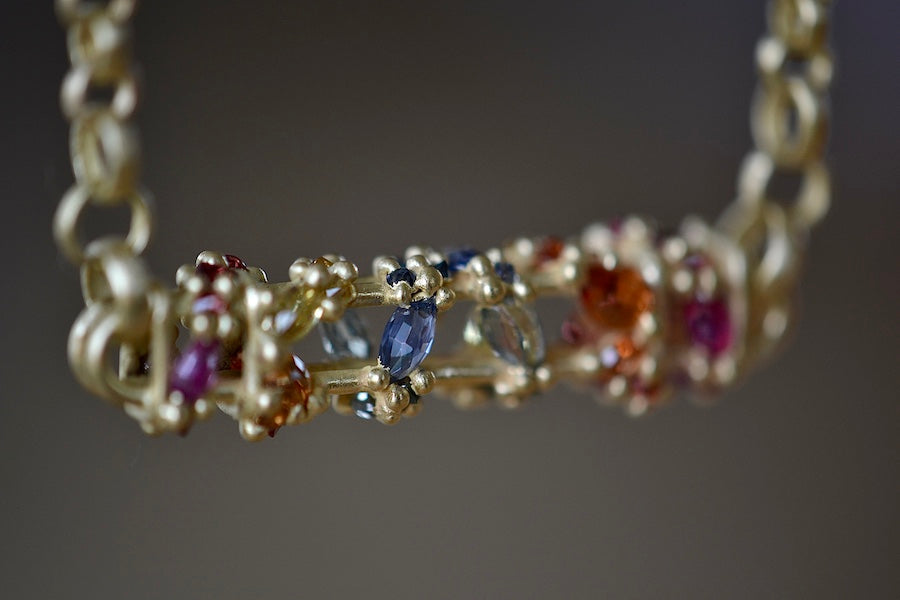 Close up of stones on Horizontal Fontaine Bar Necklace in Rainbow by Polly Wales is An oval and three dimensional bar in 18K yellow gold holds a vine of encrusted and inverted rainbow sapphires in pink, fuchsia, blue, yellow, green and red that have been cast and are accompanied by matte gold dots. Recycled Gold. Cast Not Set. Handmade in Los Angeles.