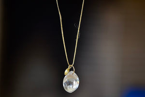 The Coconut Pendant with Seed Charm Necklace in Crystal  designed by Pippa Small is a One clear, transparent and smooth crystal stone with natural inclusions is drilled and hooked on to a gold loop, and accompanied by a bead in 18k yellow gold on a 24" golden waxed cotton cord form this necklace.