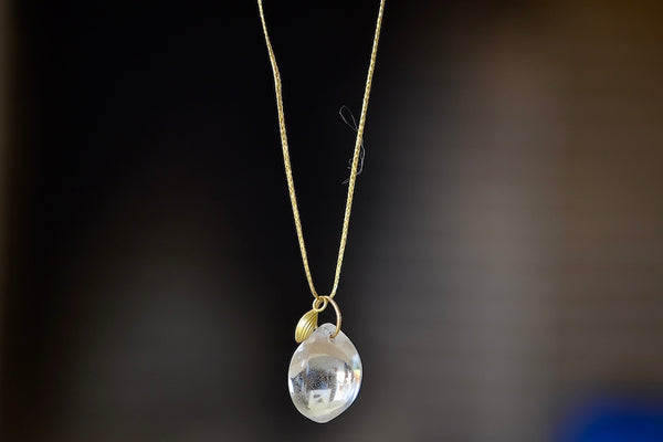 The Coconut Pendant with Seed Charm Necklace in Crystal  designed by Pippa Small is a One clear, transparent and smooth crystal stone with natural inclusions is drilled and hooked on to a gold loop, and accompanied by a bead in 18k yellow gold on a 24" golden waxed cotton cord form this necklace.