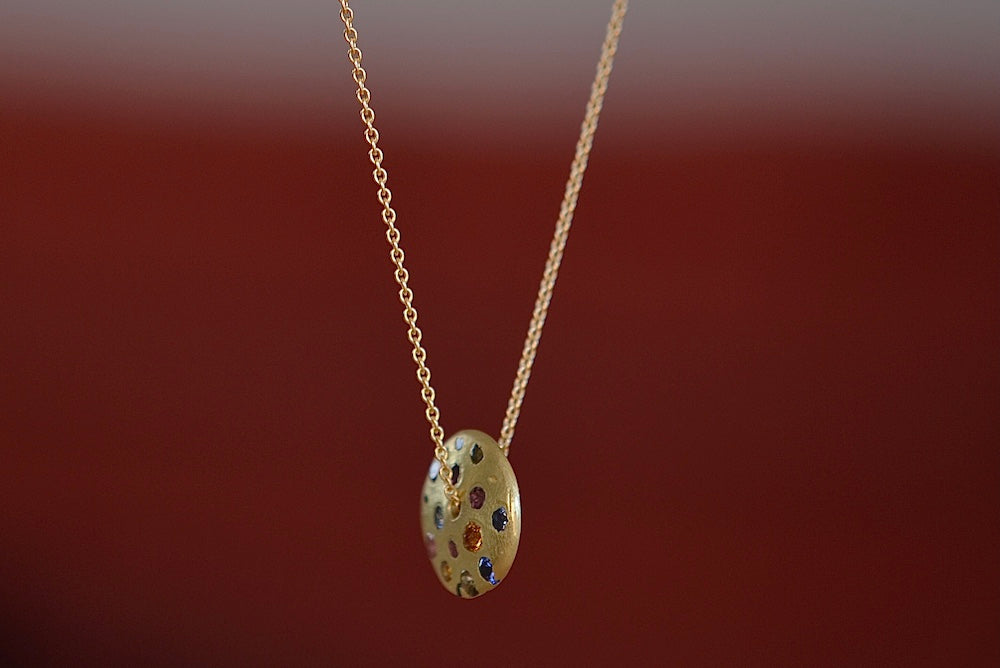 Polly Wales Spinning Disc Pendant Necklace in 18K yellow gold with scattered rainbow sapphires in orange, yellow, pink, blue, purple and lilac hangs on a beautiful chain 22" cast in place and cast not set in recycled gold.